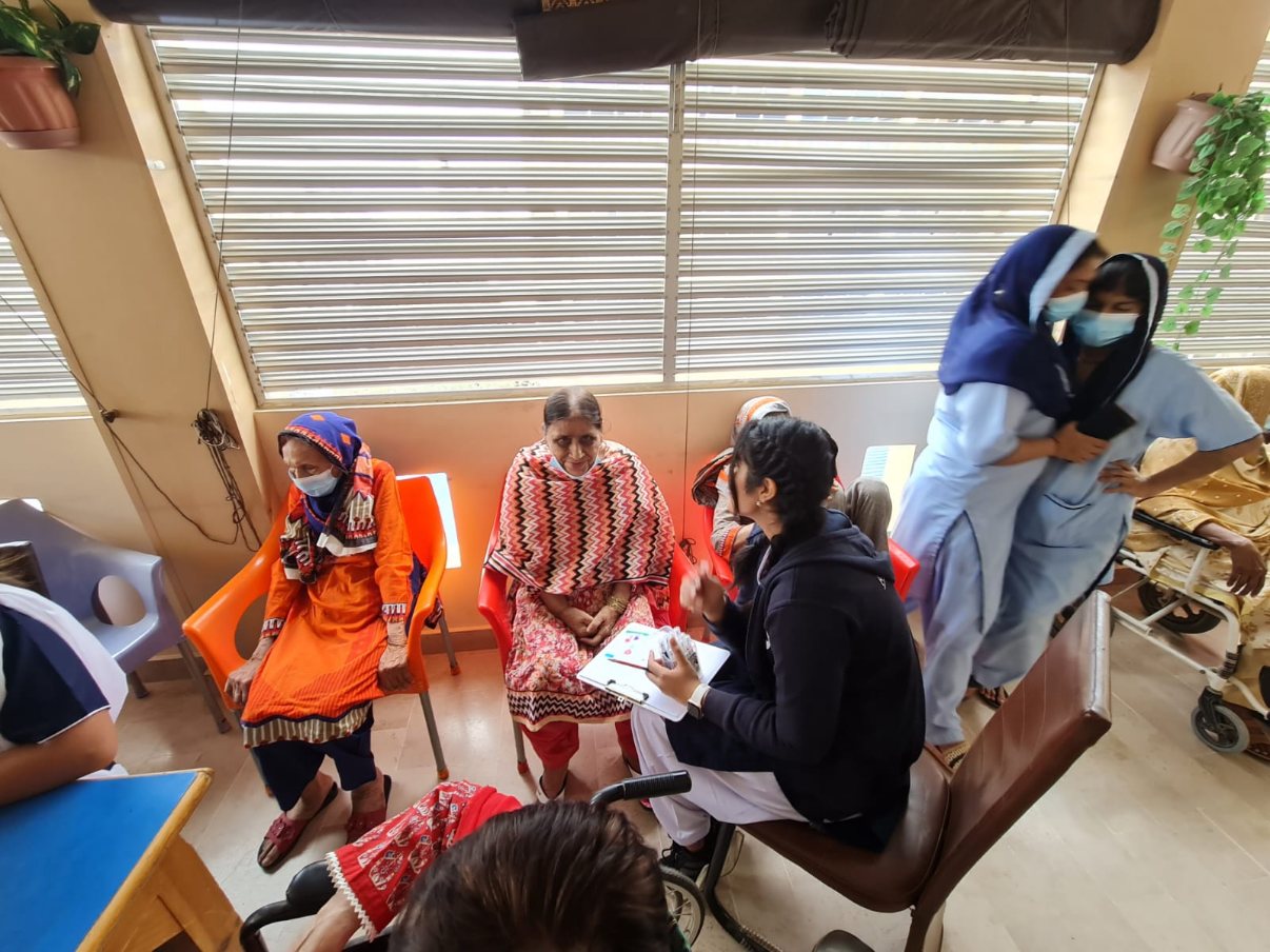 Residents seated in a row of orange and red chairs beneath a window, with a volunteer holding a clipboard
