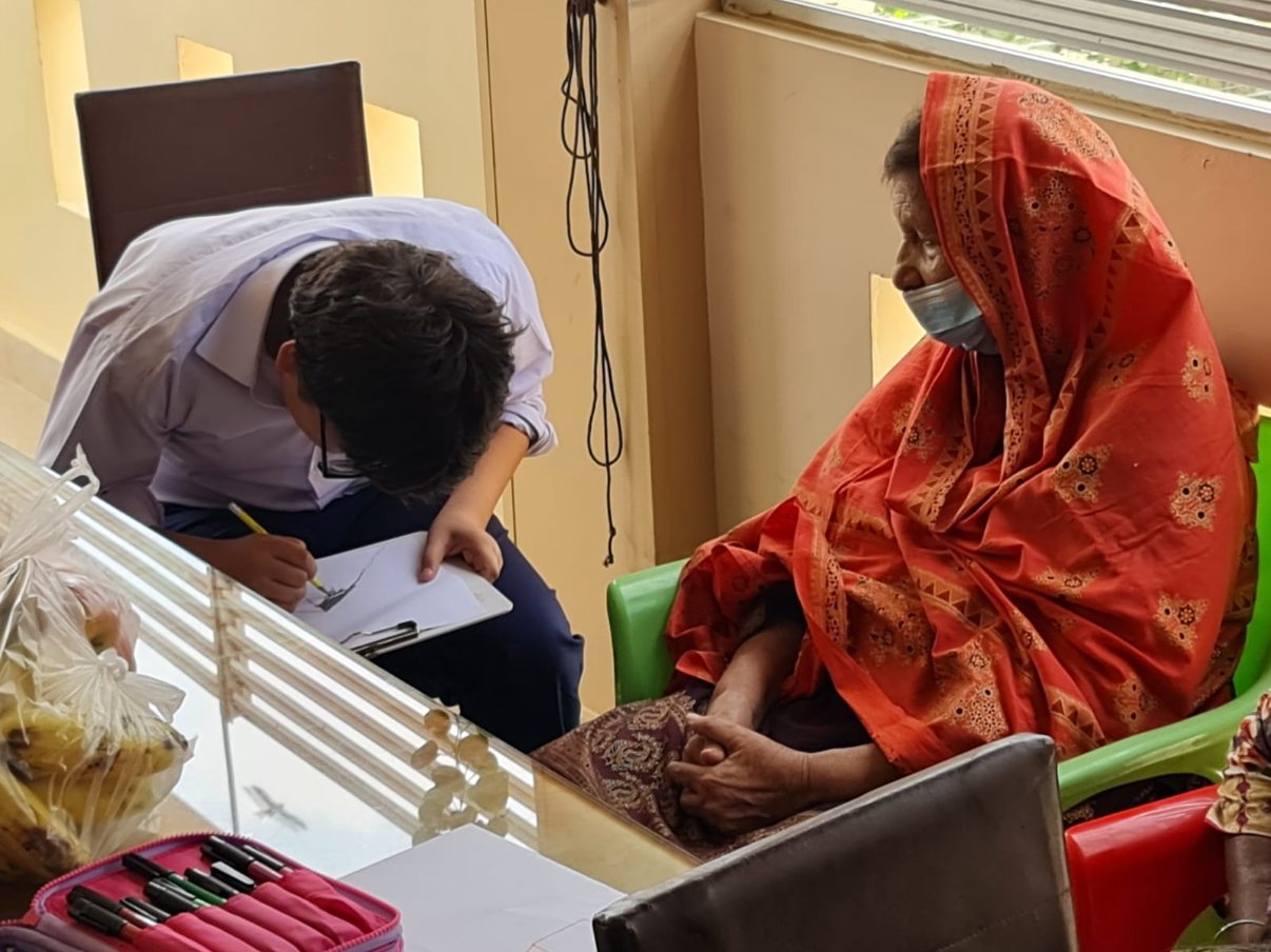 A student leaning over a clipboard to write beside a resident wrapped in an orange shawl