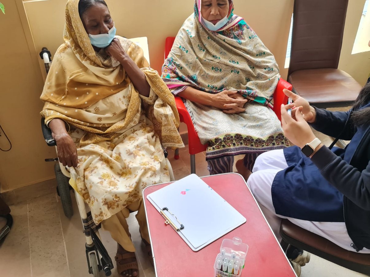 Two residents seated beside a table with a clipboard and coloured markers while a volunteer gestures as she explains something