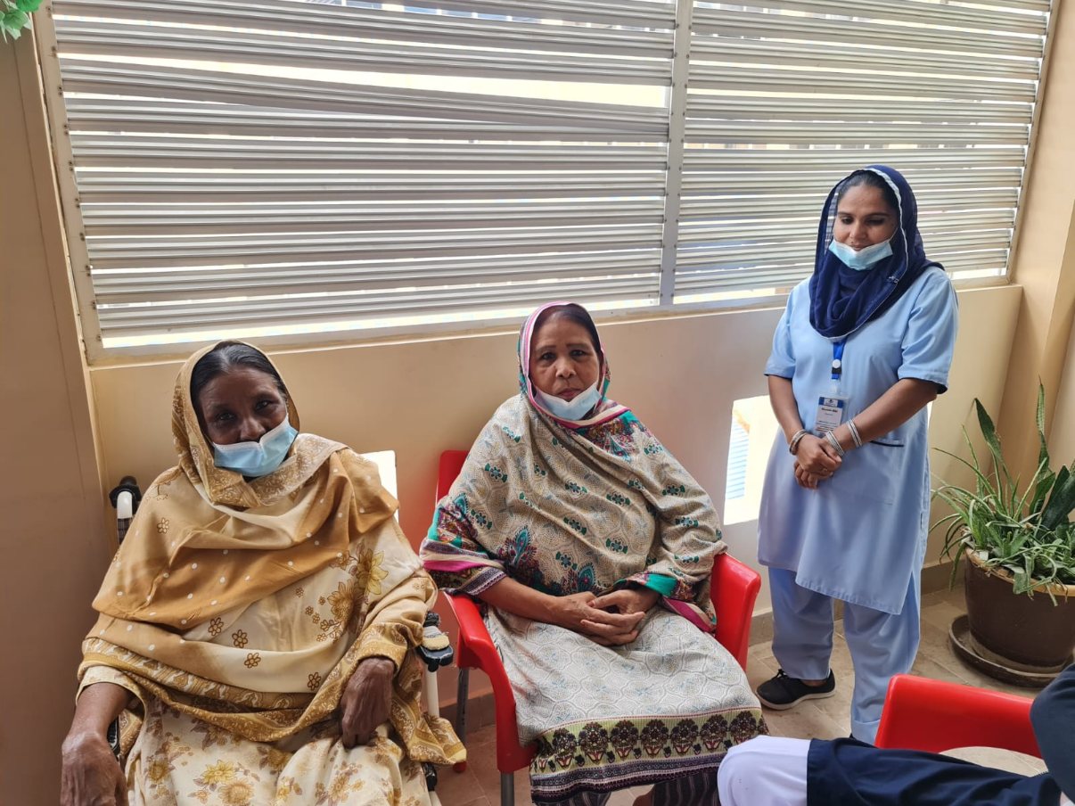 Two residents seated on red chairs by a window, one in a wheelchair, with a caregiver in light blue standing beside them