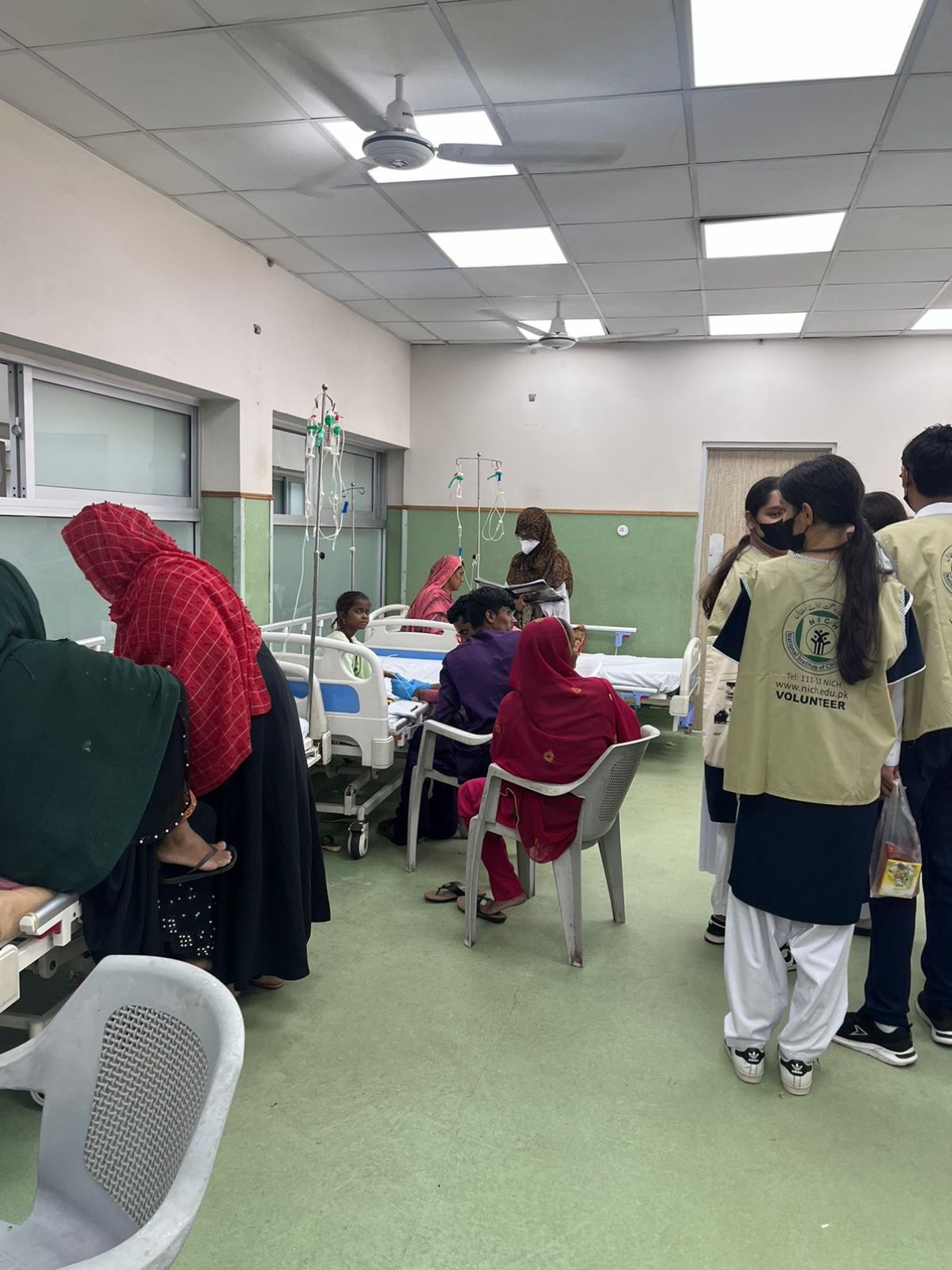 Student volunteers in vests standing in a hospital ward with beds and drip stands while families sit nearby