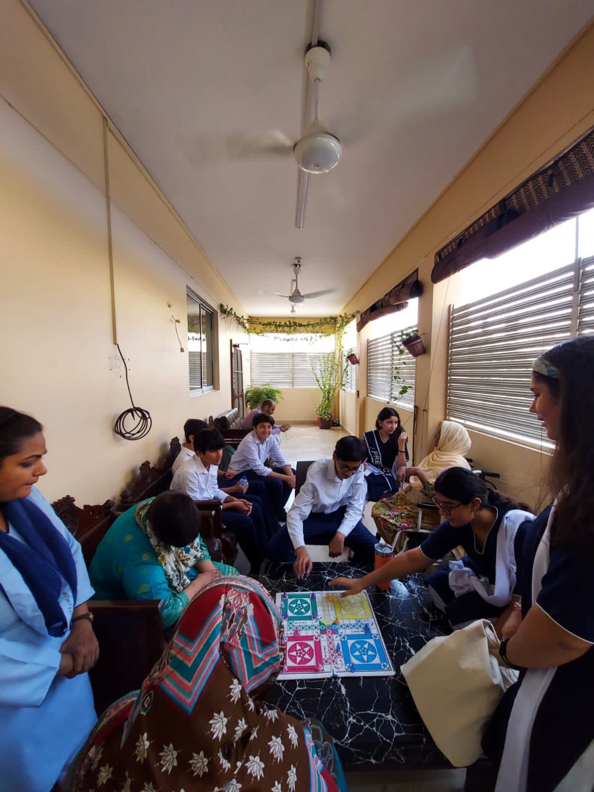 A group playing Ludo on a table in a bright corridor with residents, caregivers and student volunteers